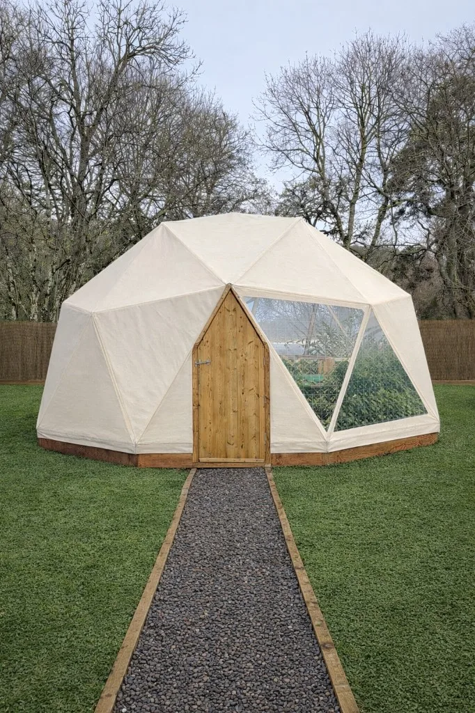 Geodesic dome with light canvas cover, timber door and triangular viewing windows showing plants inside, pebble path and lawn